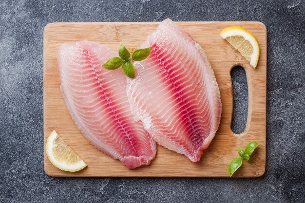 Two fish fillets on a wooden cutting board with lemon slices and basil.