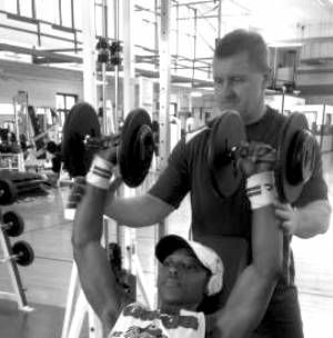 A man is helping another man lift dumbbells in a gym.