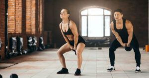 A man and a woman are squatting with kettlebells in a gym.