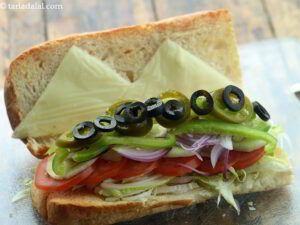 A sub sandwich with cheese , tomatoes , cucumbers , onions and olives on a table.