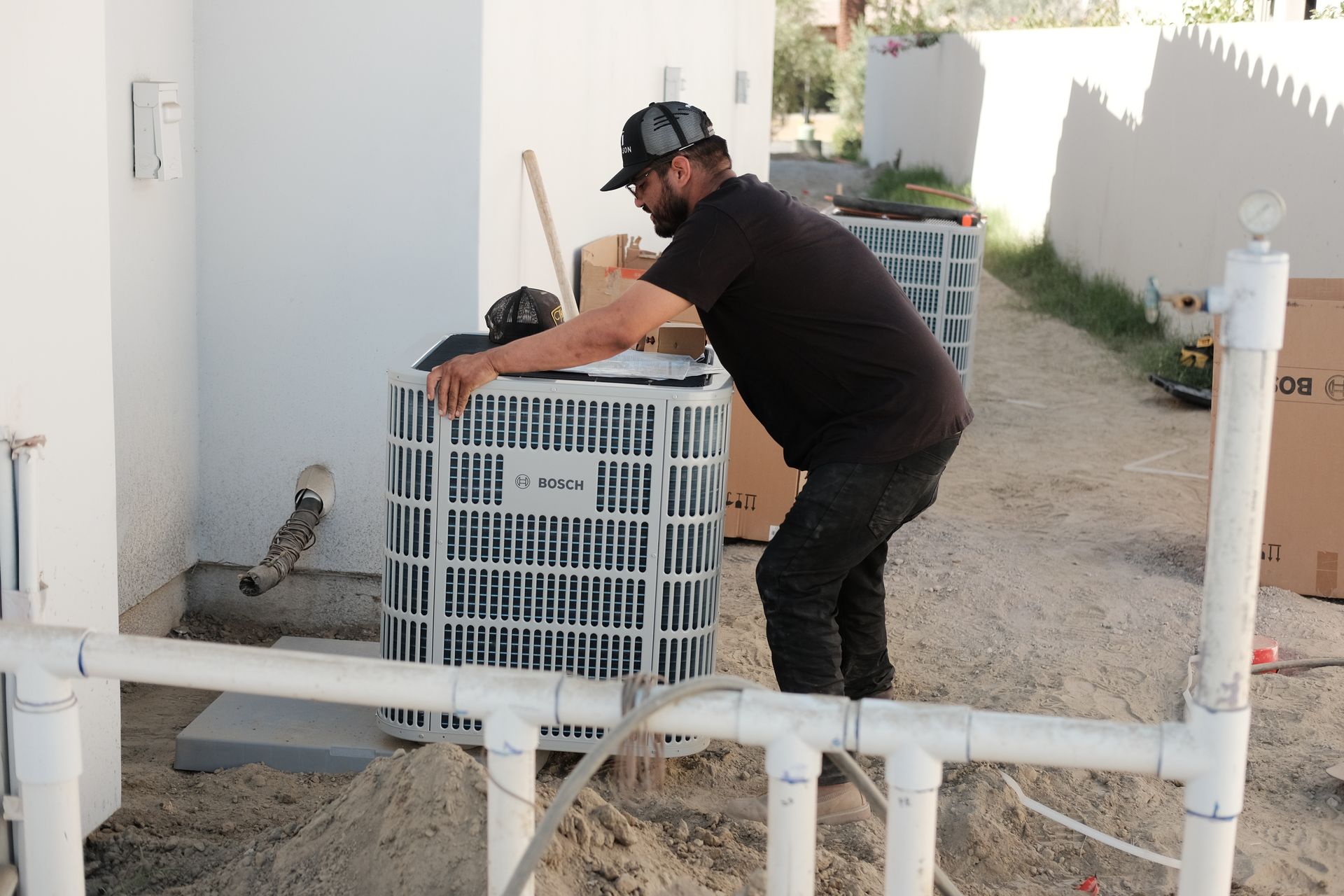 A person in black clothing working on an air conditioning unit outside near a white wall.