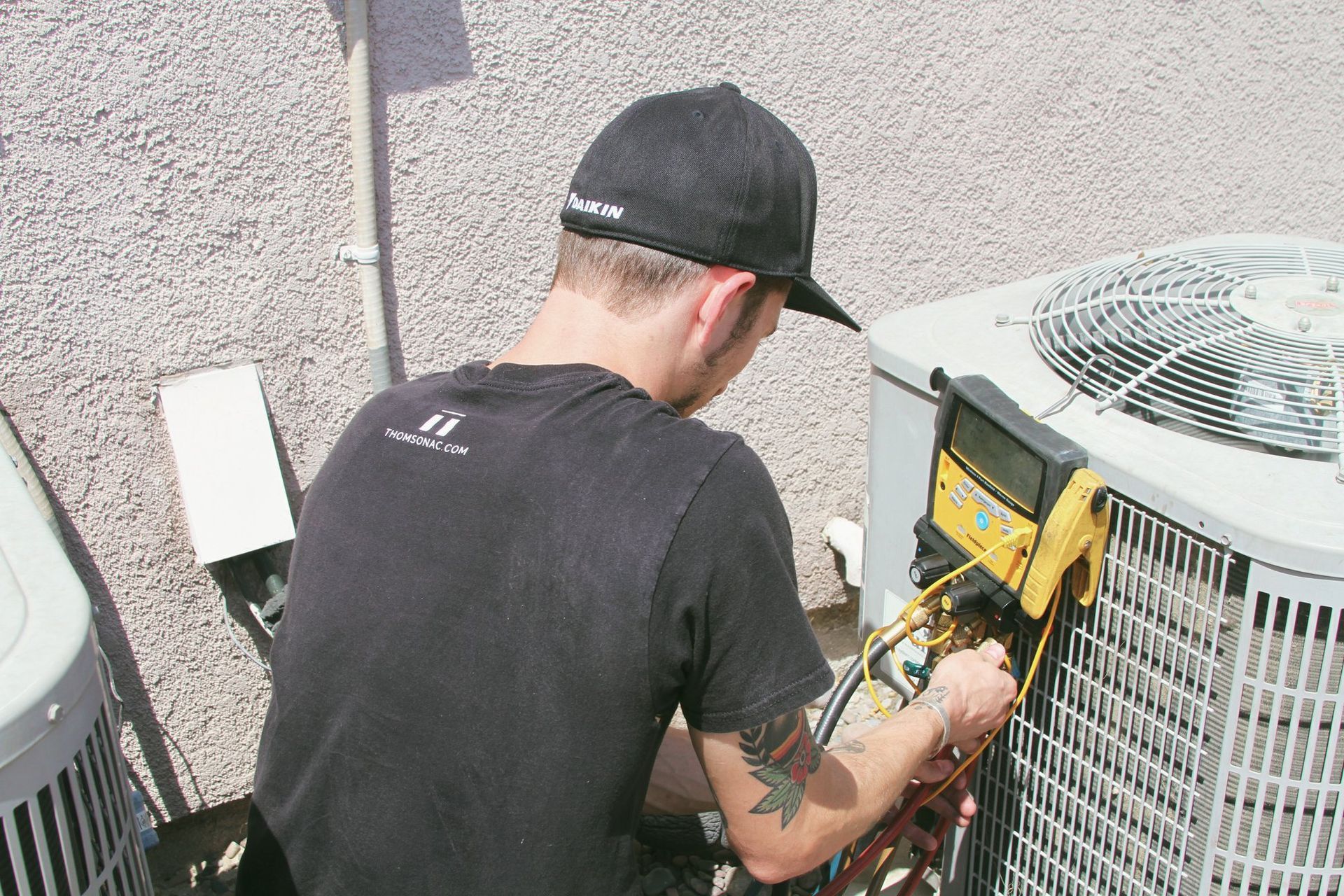 HVAC technician in black cap and t-shirt, working on an outdoor air conditioning unit with gauges, by a stucco wall.