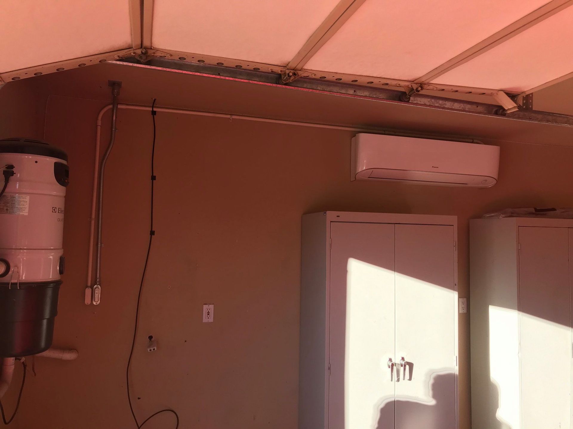 An air conditioner and central vacuum system mounted on a garage wall with cabinets and overhead beams.