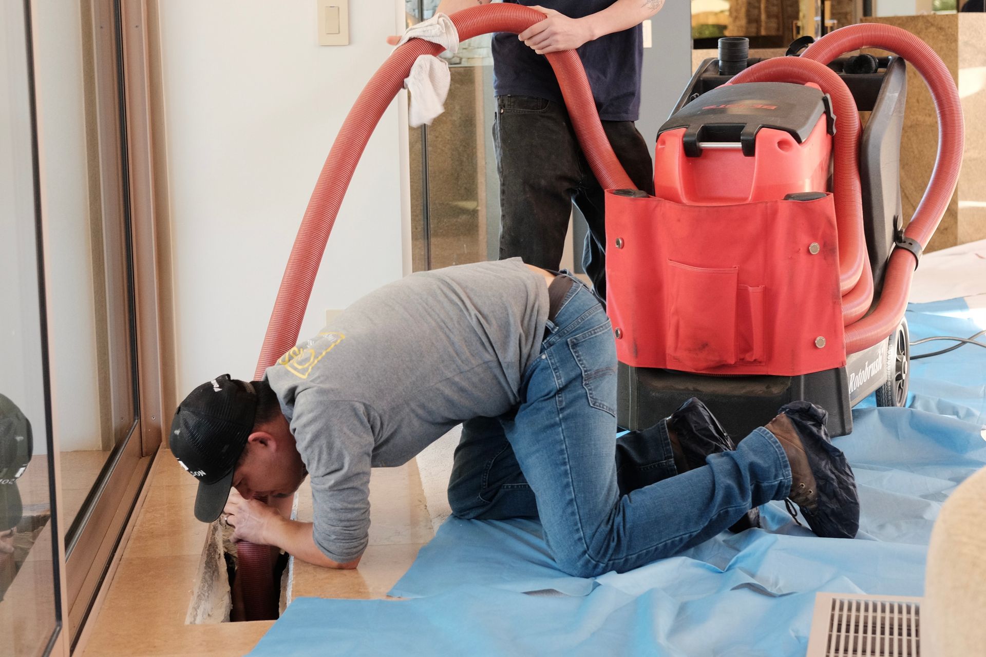 Two people using a red machine to clean an opening in a floor; one leans in, the other holds a hose.