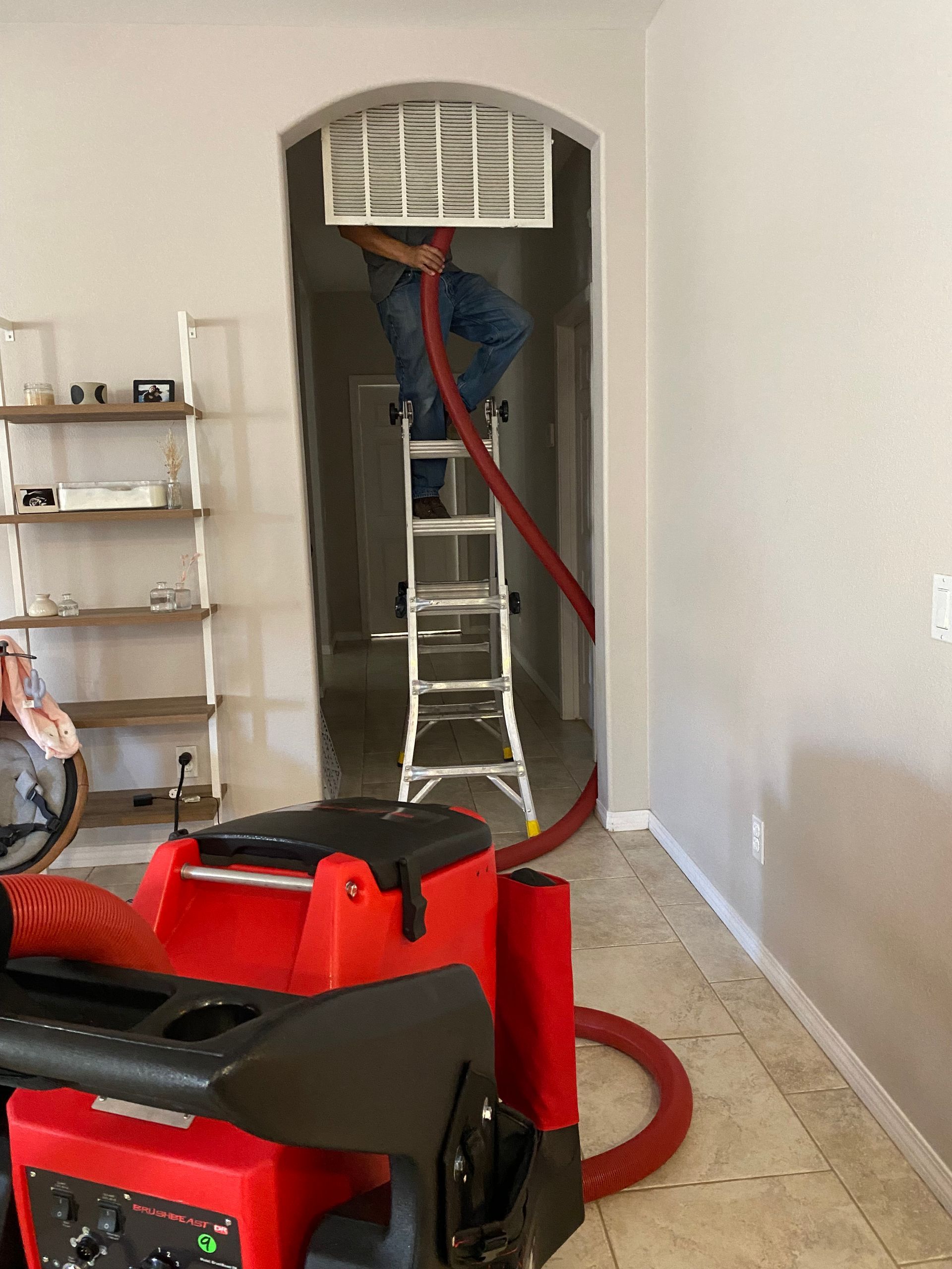 Person on ladder cleaning an air vent with a red hose connected to a red machine in a hallway.