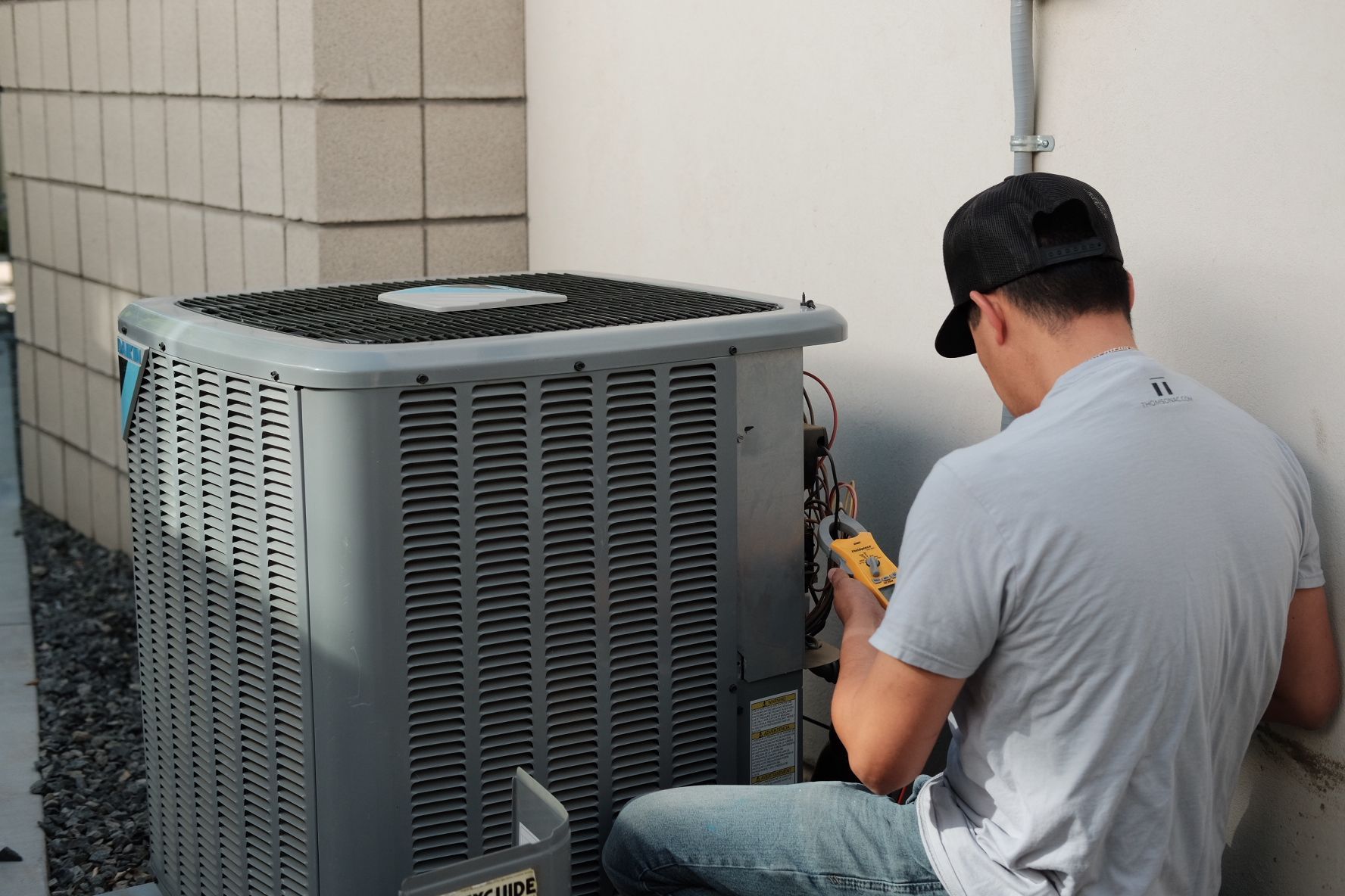 HVAC technician kneeling, inspecting an air conditioning unit. Gray unit, white wall, brick block, holding a tool.