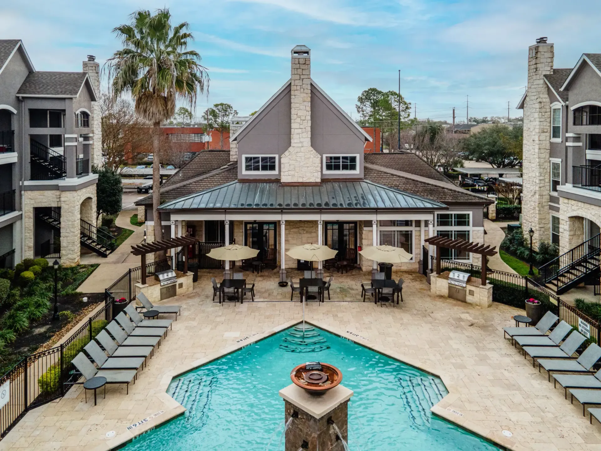 Aerial view of a pool, patio with tables and umbrellas, and apartment buildings on a sunny day.