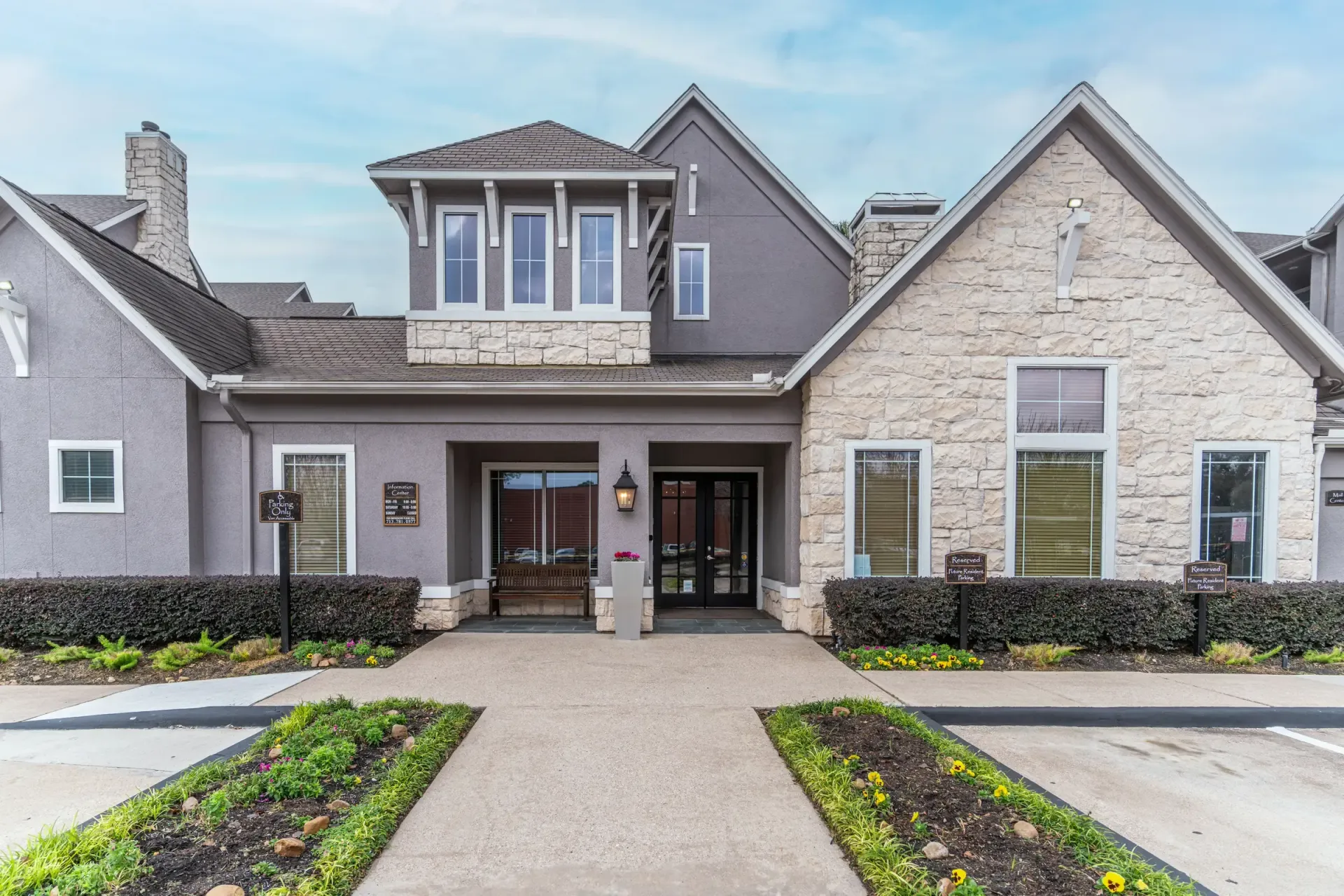 Stone and gray building with a central entrance, windows, and a brown roof. Landscaping in the foreground.