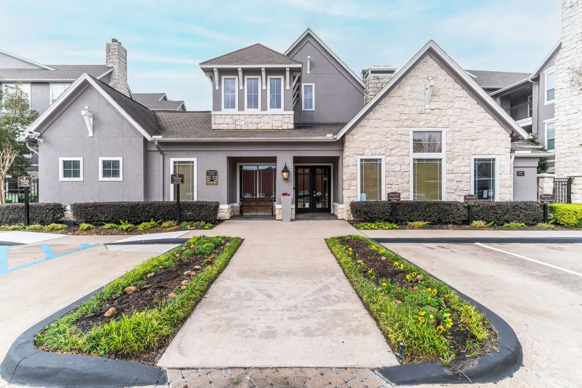 Exterior of a building with stone and grey siding. Entrance with pathway and landscaping. Parking spaces in front.