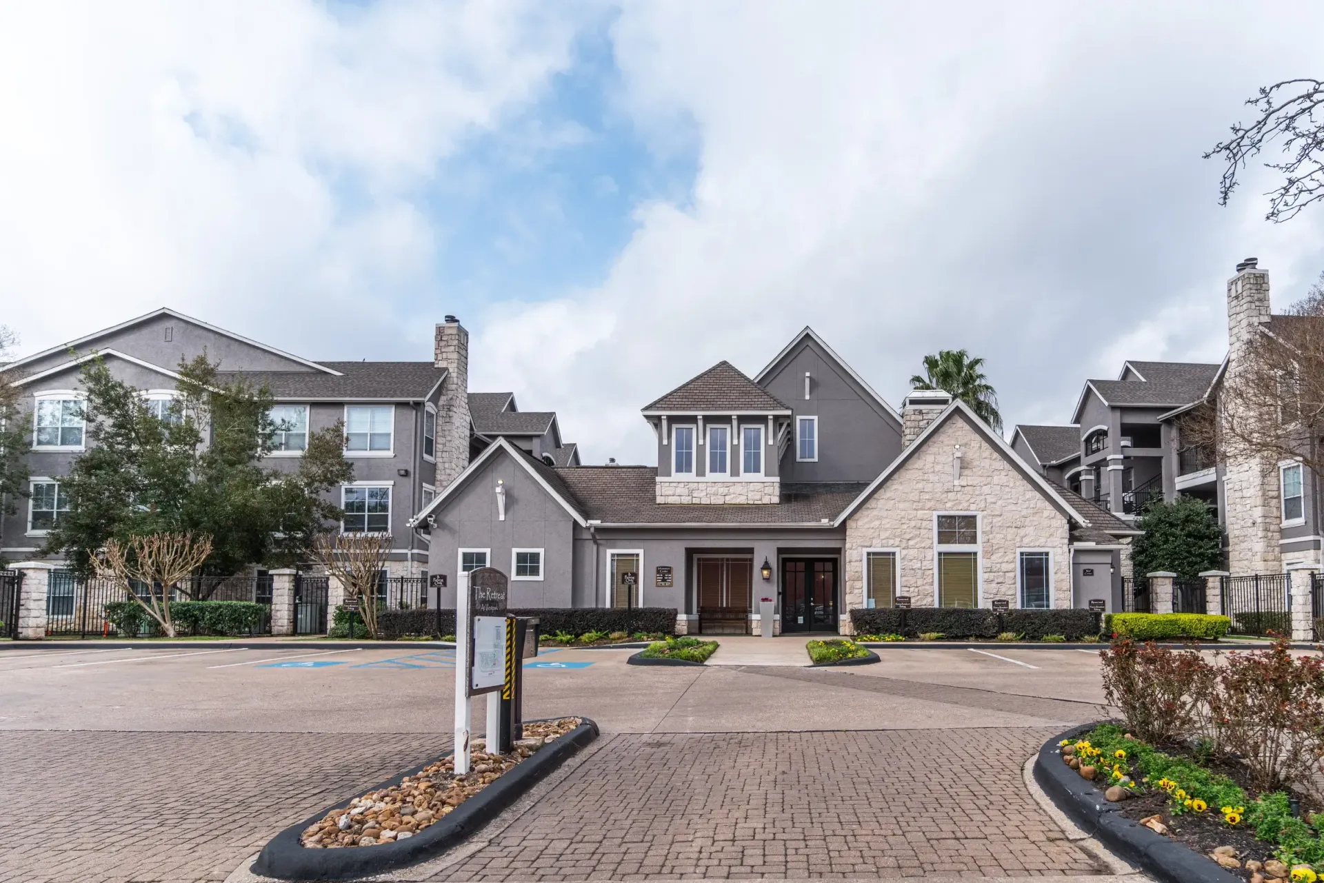 Exterior of a gray building with stone accents and a brick-paved driveway under a cloudy sky.