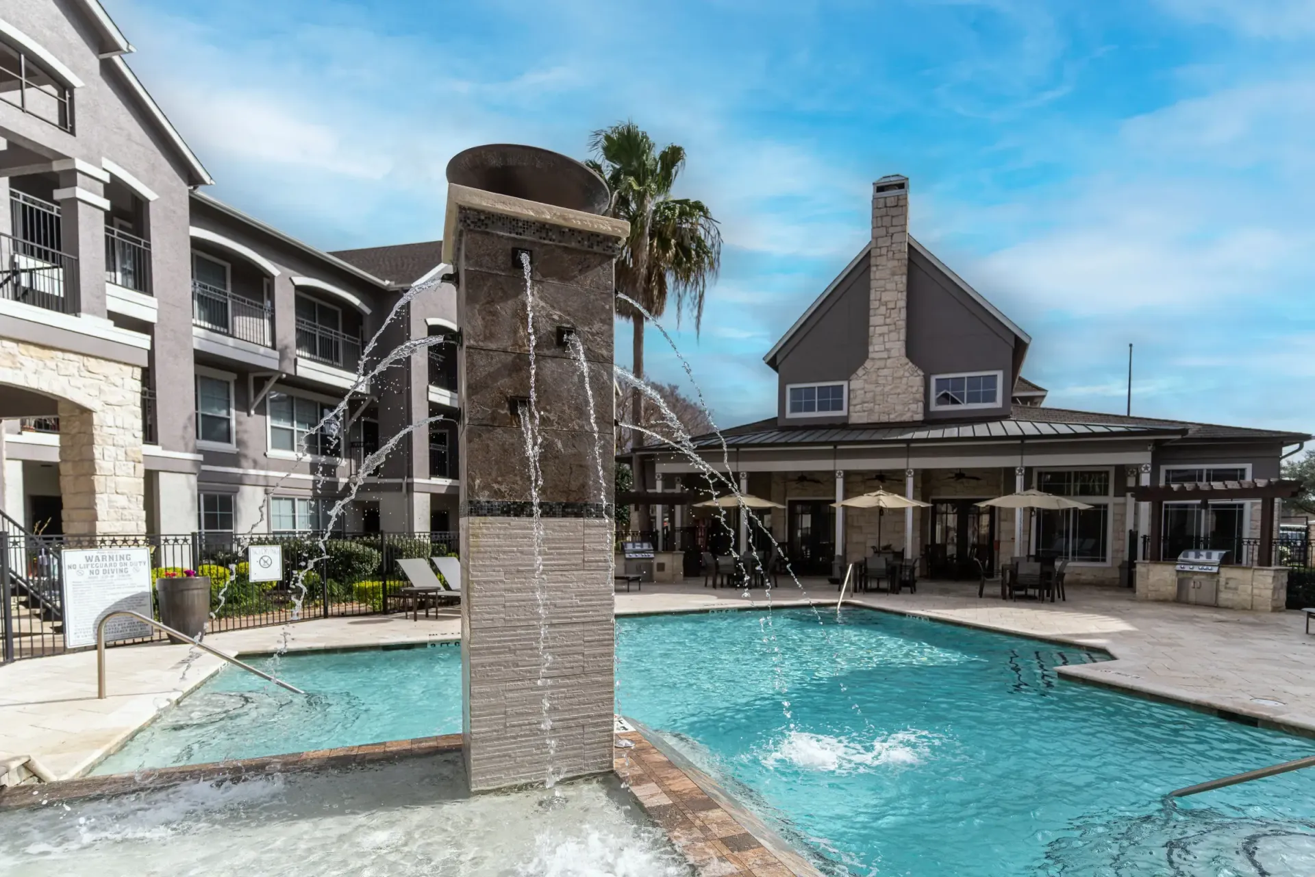 Swimming pool with a water feature, lounge chairs, and buildings under a blue sky.