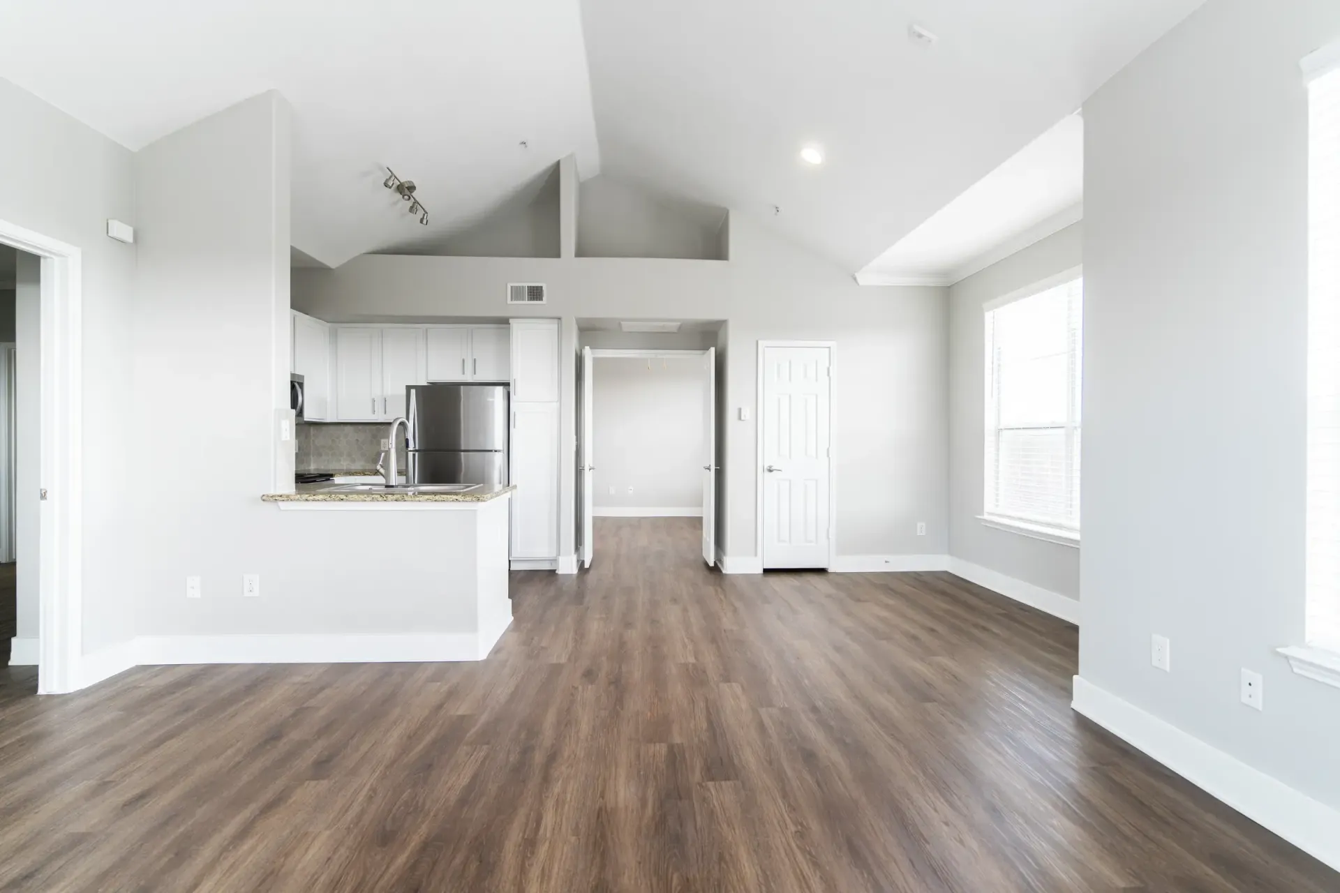 Spacious, empty living area with wood-look flooring, white walls, and a vaulted ceiling. Kitchen and hallway visible.