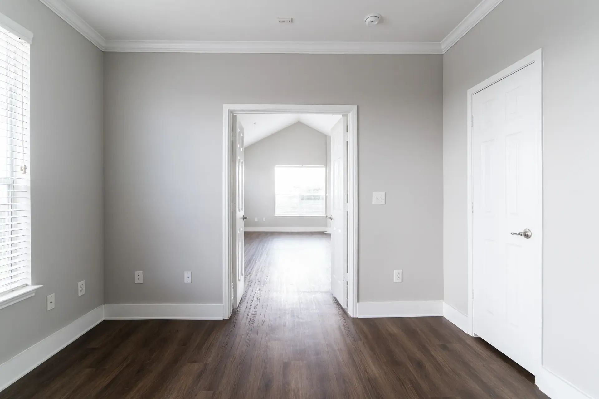 Empty room with gray walls, white trim, and dark wood-look flooring. Doorway leads to another room with window.