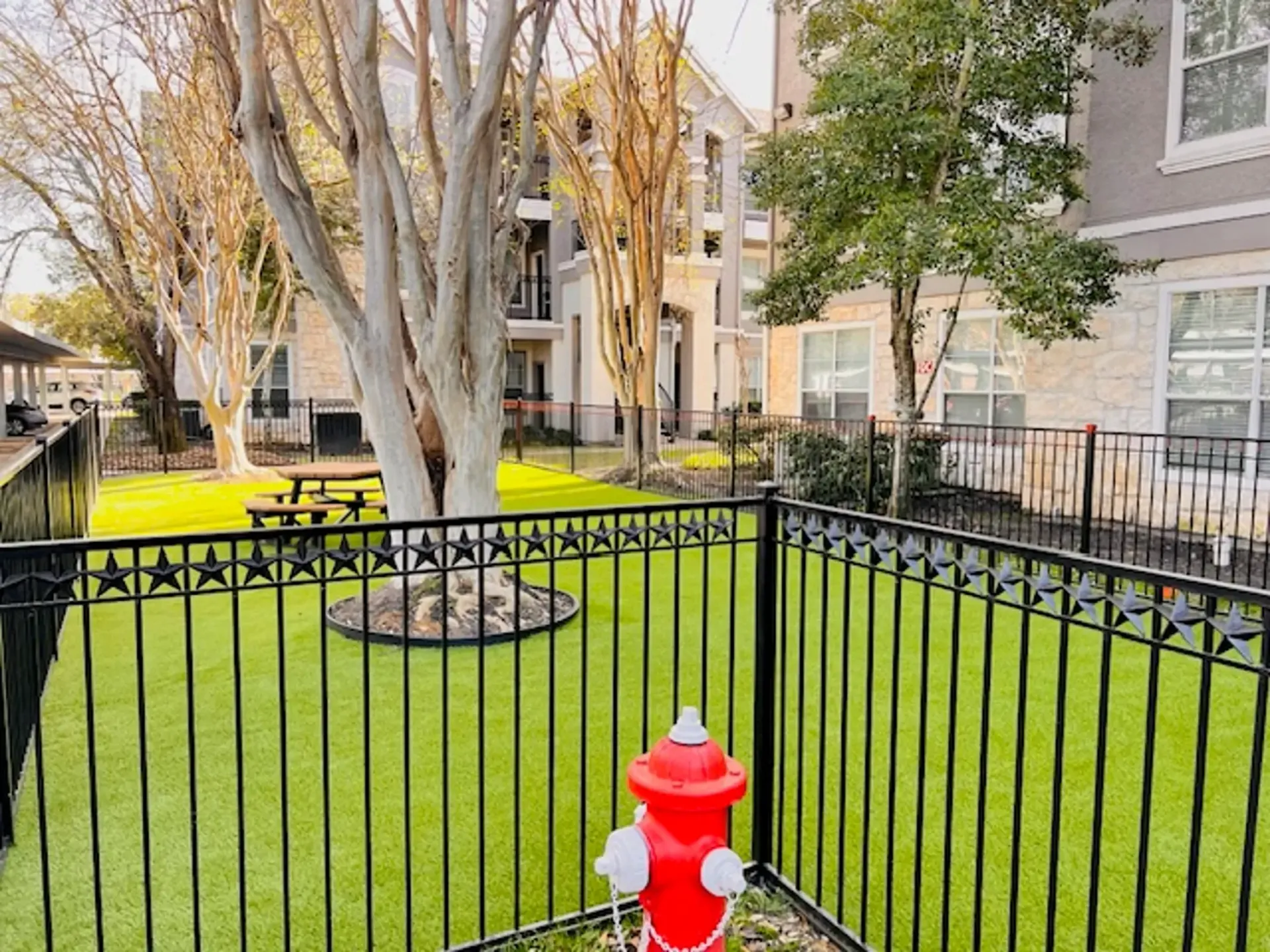 A fenced dog park with green turf, a red fire hydrant, and apartment buildings in the background.