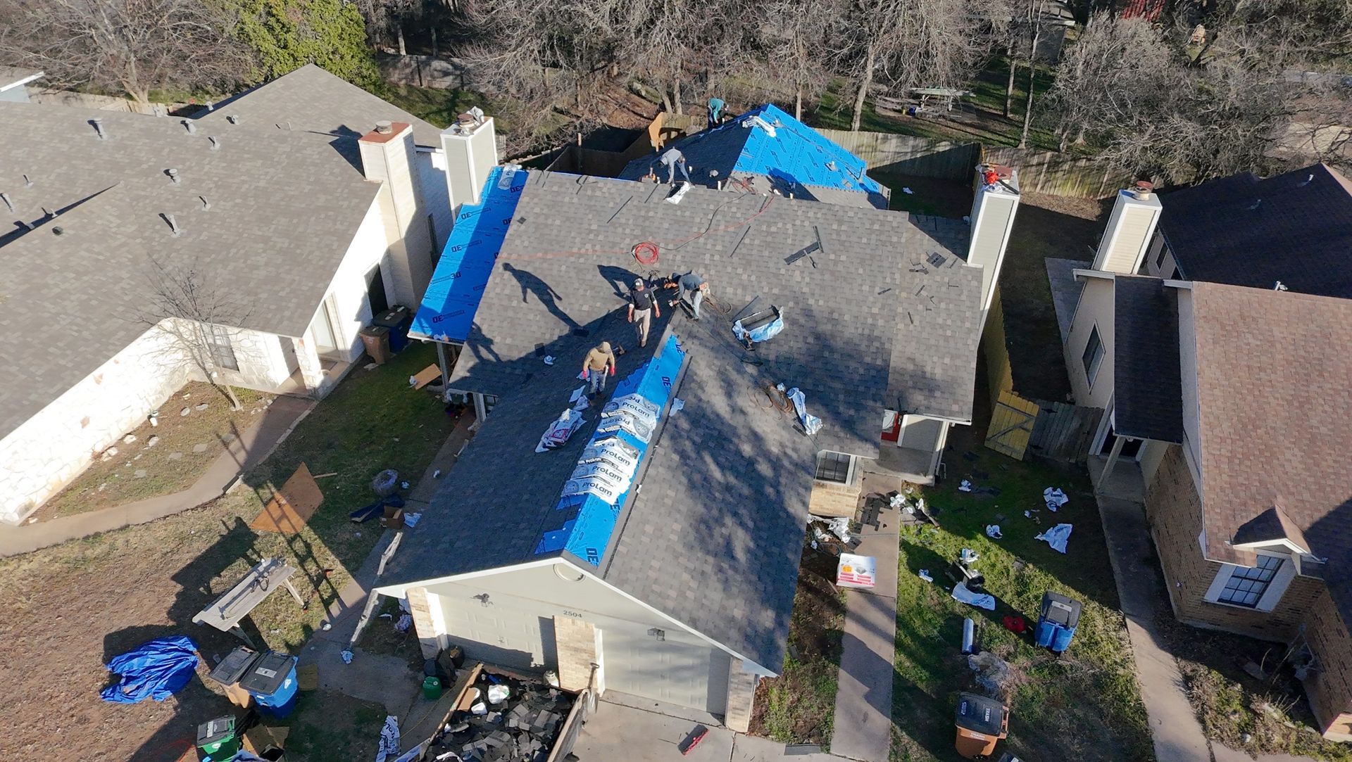 Aerial view of a house roof covered with blue tarps after storm damage, with debris around the yard.