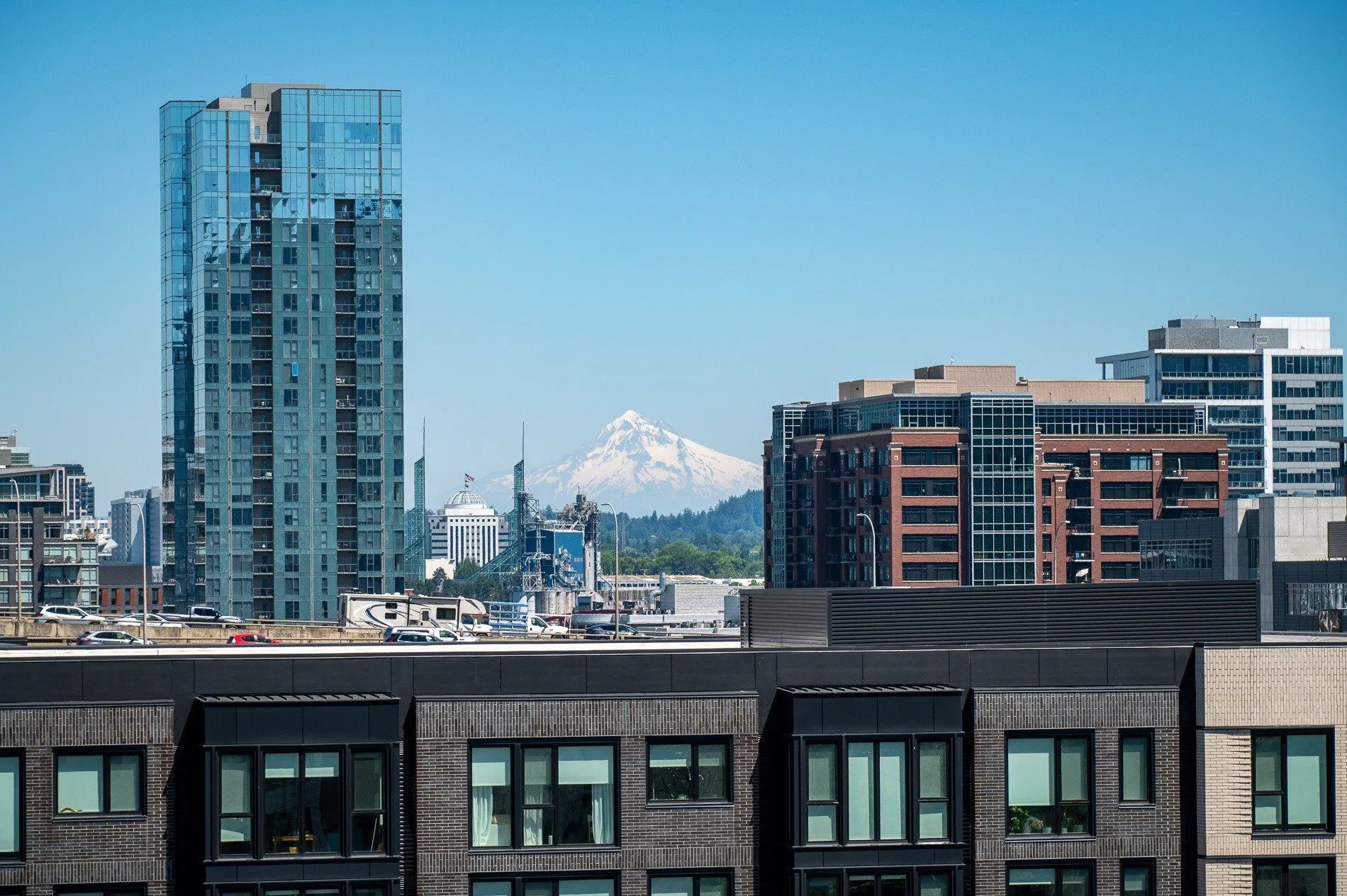 City skyline with a tall, blue glass building and snow-capped mountain in the background under a blue sky.