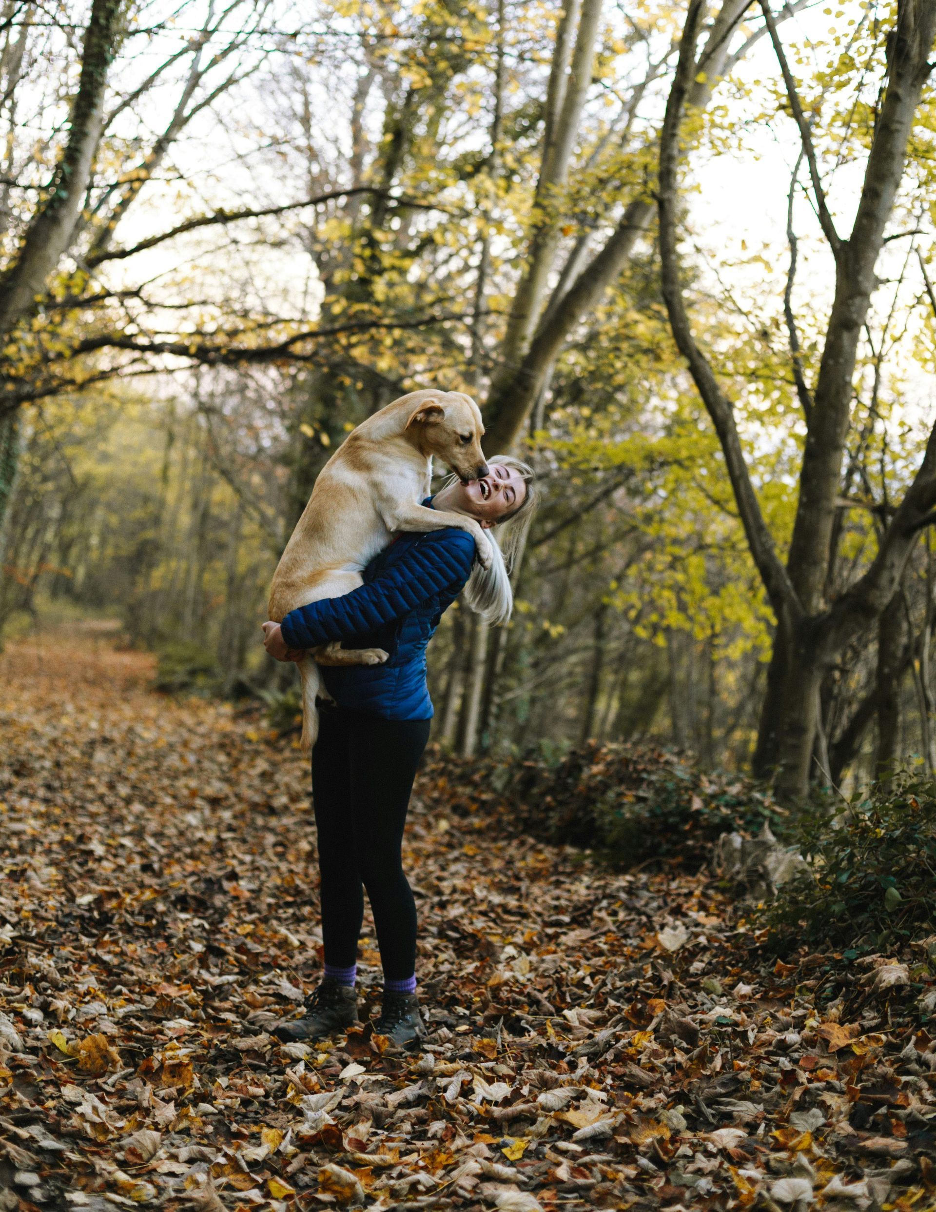 Person holding a large, light-colored dog in a forest path with fall leaves. The dog is nuzzling the person’s face.