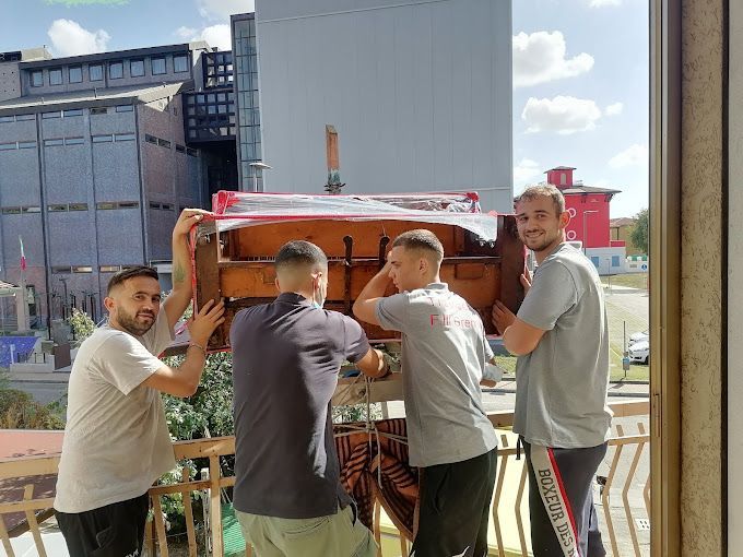Quattro uomini trasportano un armadio di legno rosso su un balcone esterno, con degli edifici sullo sfondo.
