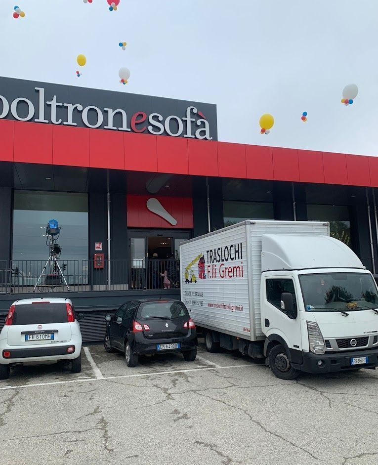 Vetrina di un negozio di mobili con camion per le consegne e auto parcheggiate sotto un cielo nuvoloso.