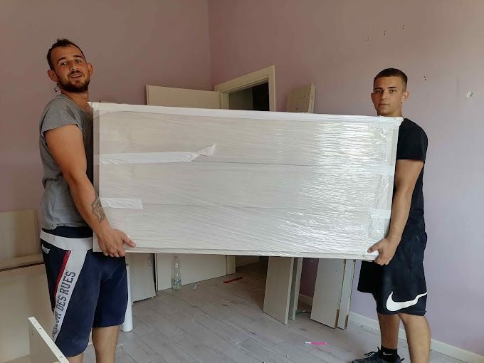 Due uomini trasportano un grande materasso imballato attraverso una stanza non ancora finita