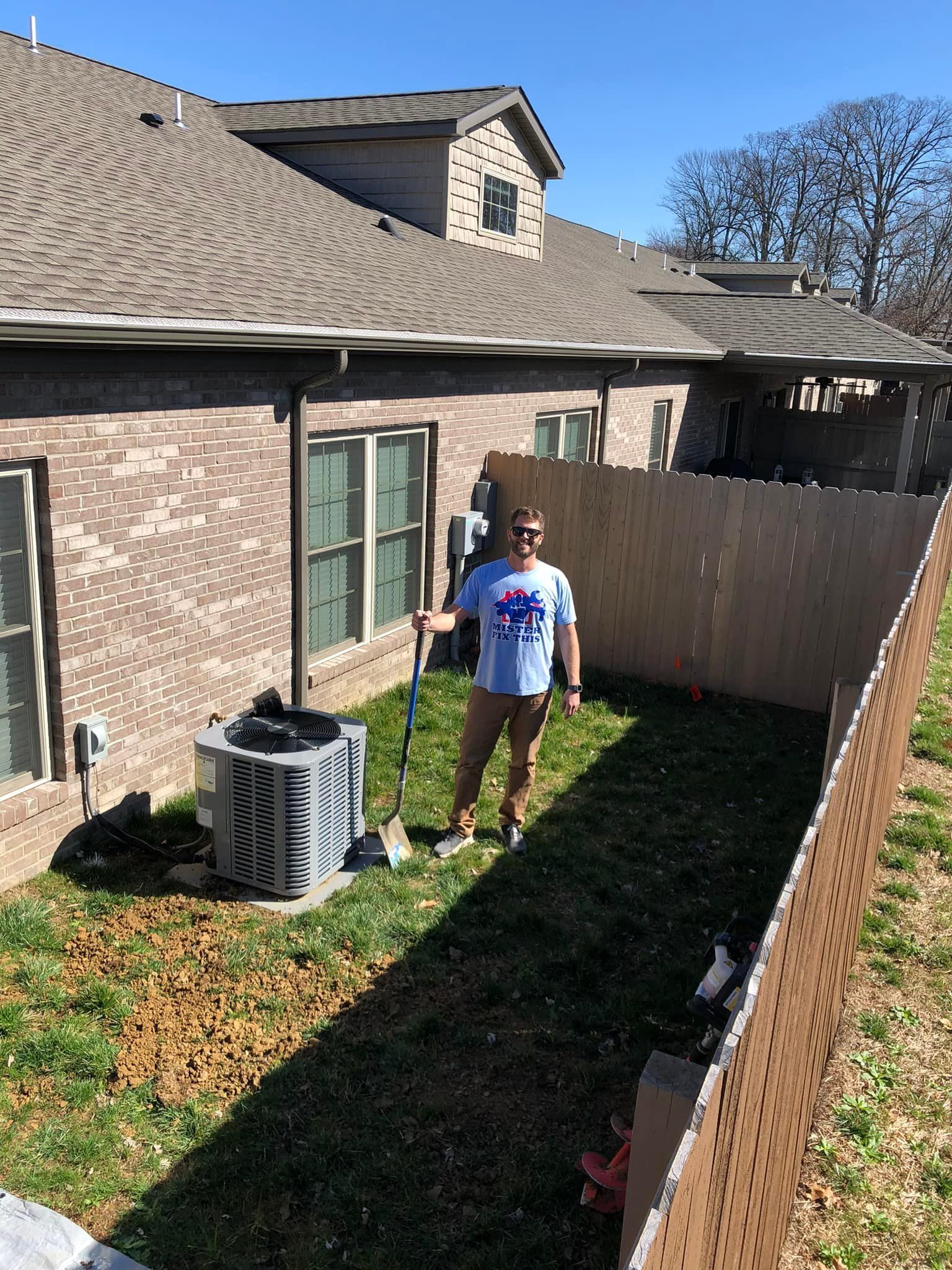 Man raking leaves in a small yard between a brick house and wooden fence on a sunny day.