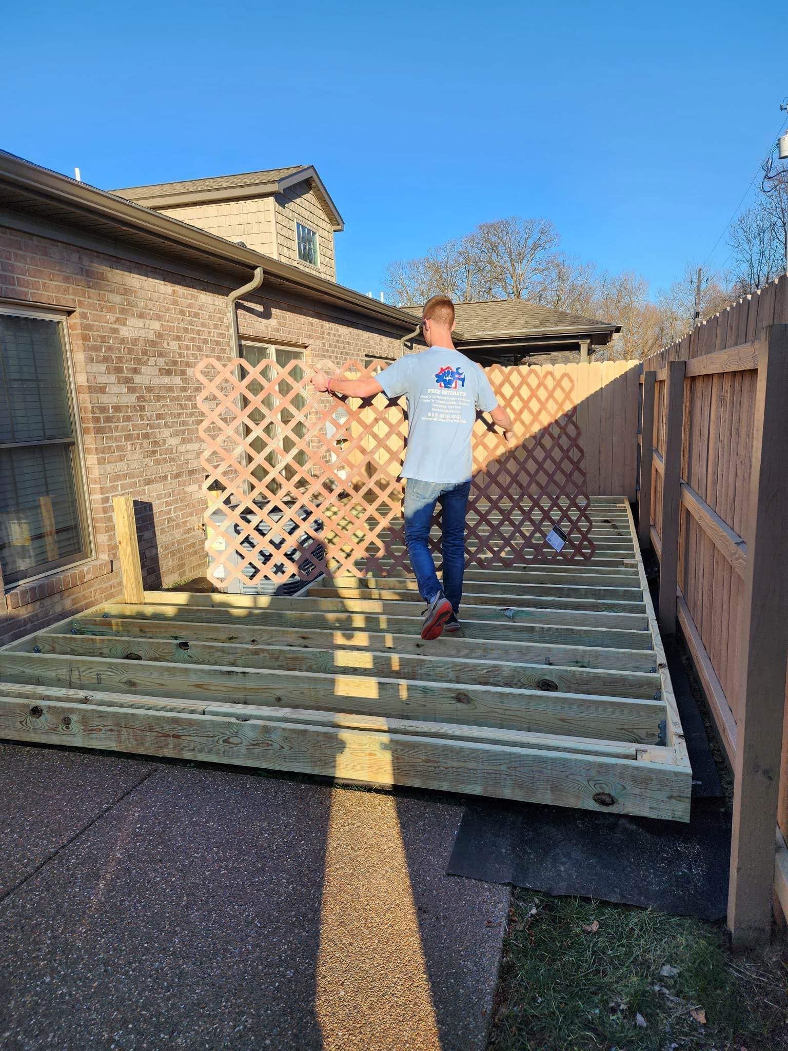 Person standing on a partially built wooden deck, arms outstretched. Lattice screen and fence in the background, blue sky.