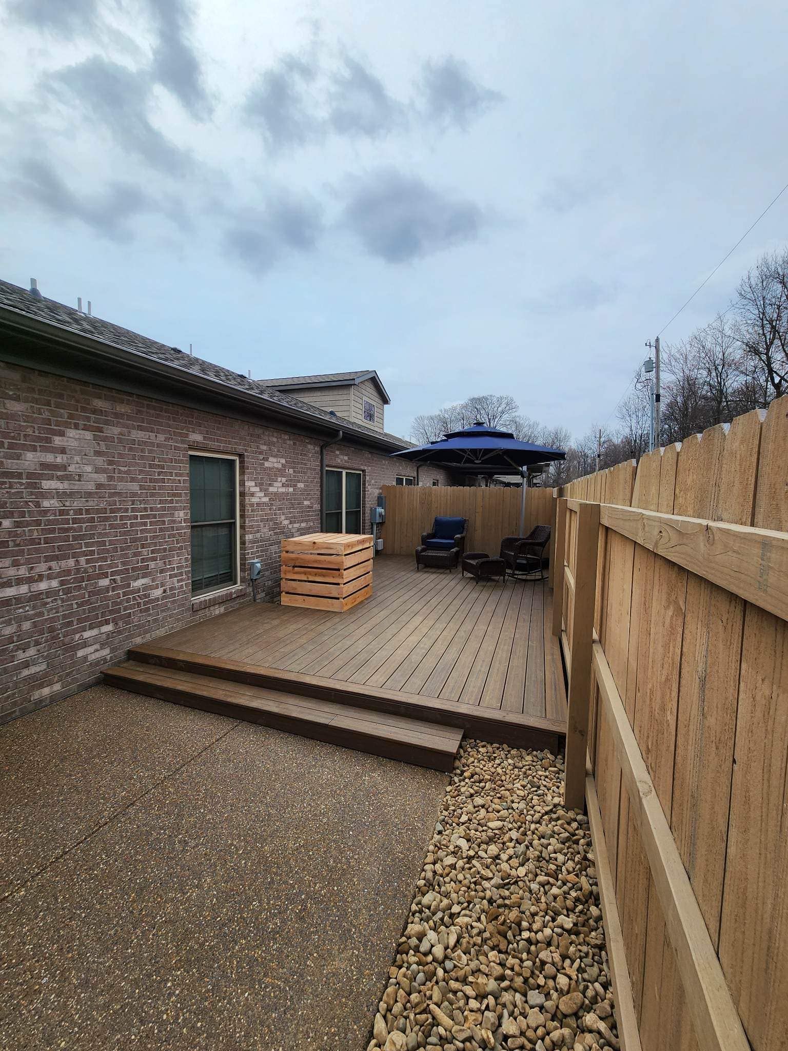 Backyard with wooden deck and brick house next to a wooden fence. Gravel and cloudy sky.