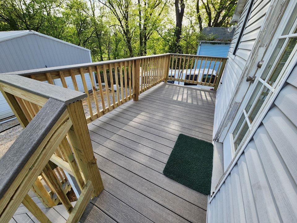 Wooden deck with railing attached to a light-colored house. Green trees in the background.