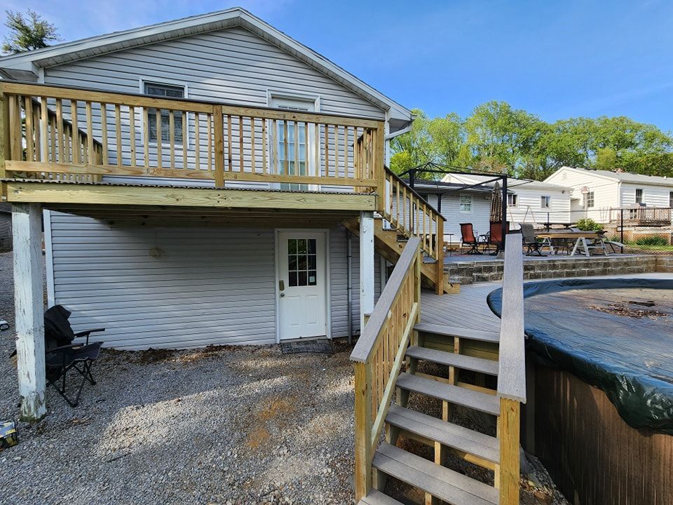 House with wooden deck and stairs leading to a pool area. Gray siding, gravel ground.