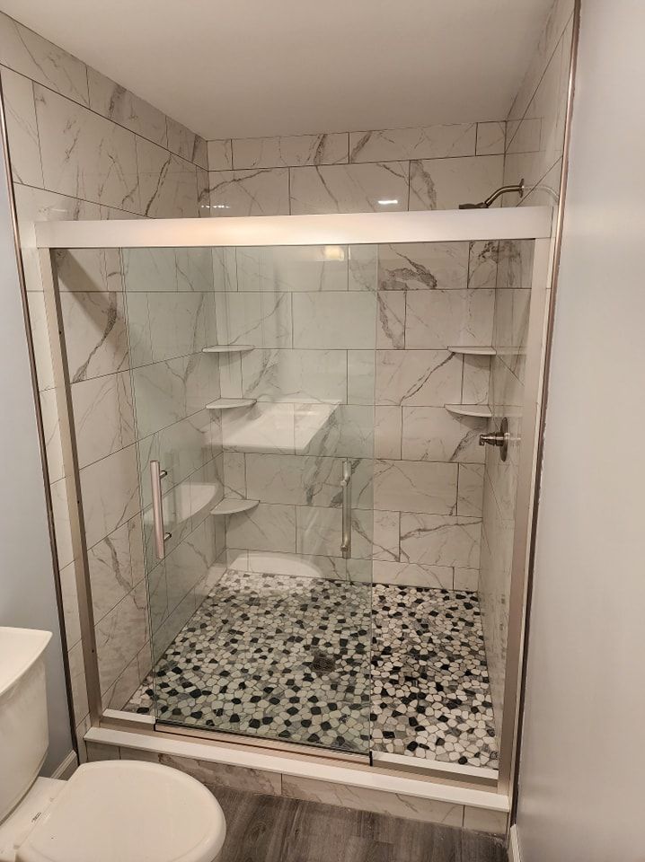 Shower with clear glass doors, light-colored marble-look tile, pebble floor, and built-in shelves.