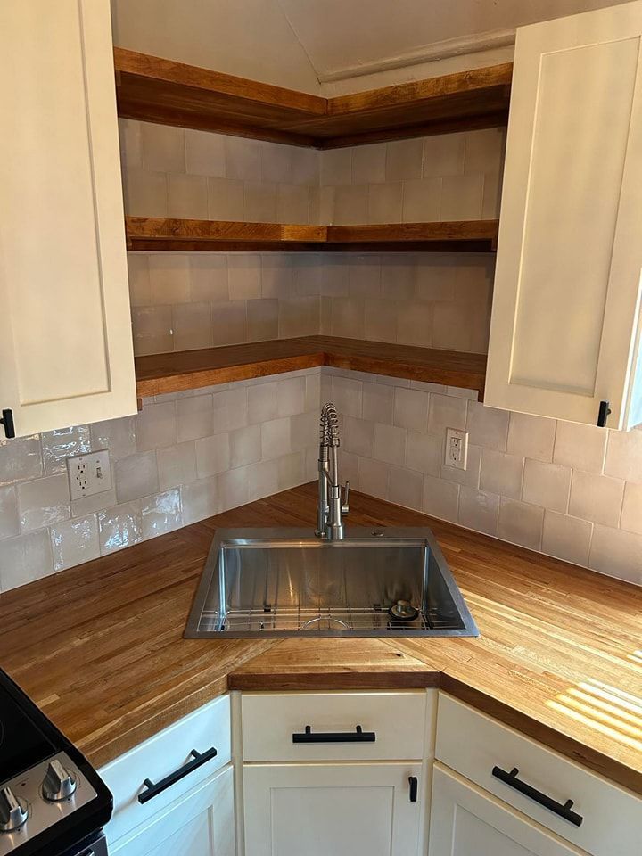 Corner kitchen sink with wooden countertops and open shelving, white cabinets, and white tiled backsplash.