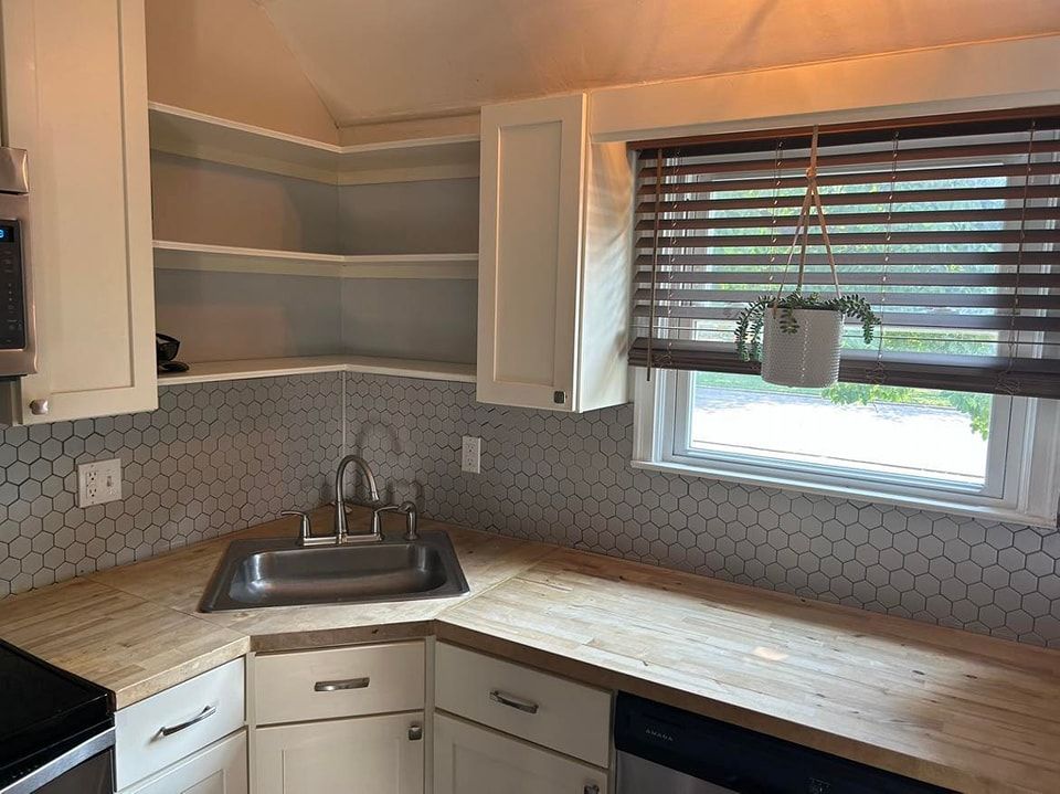 Kitchen with white cabinets, wood countertop, hexagon tile backsplash, and window with blinds.