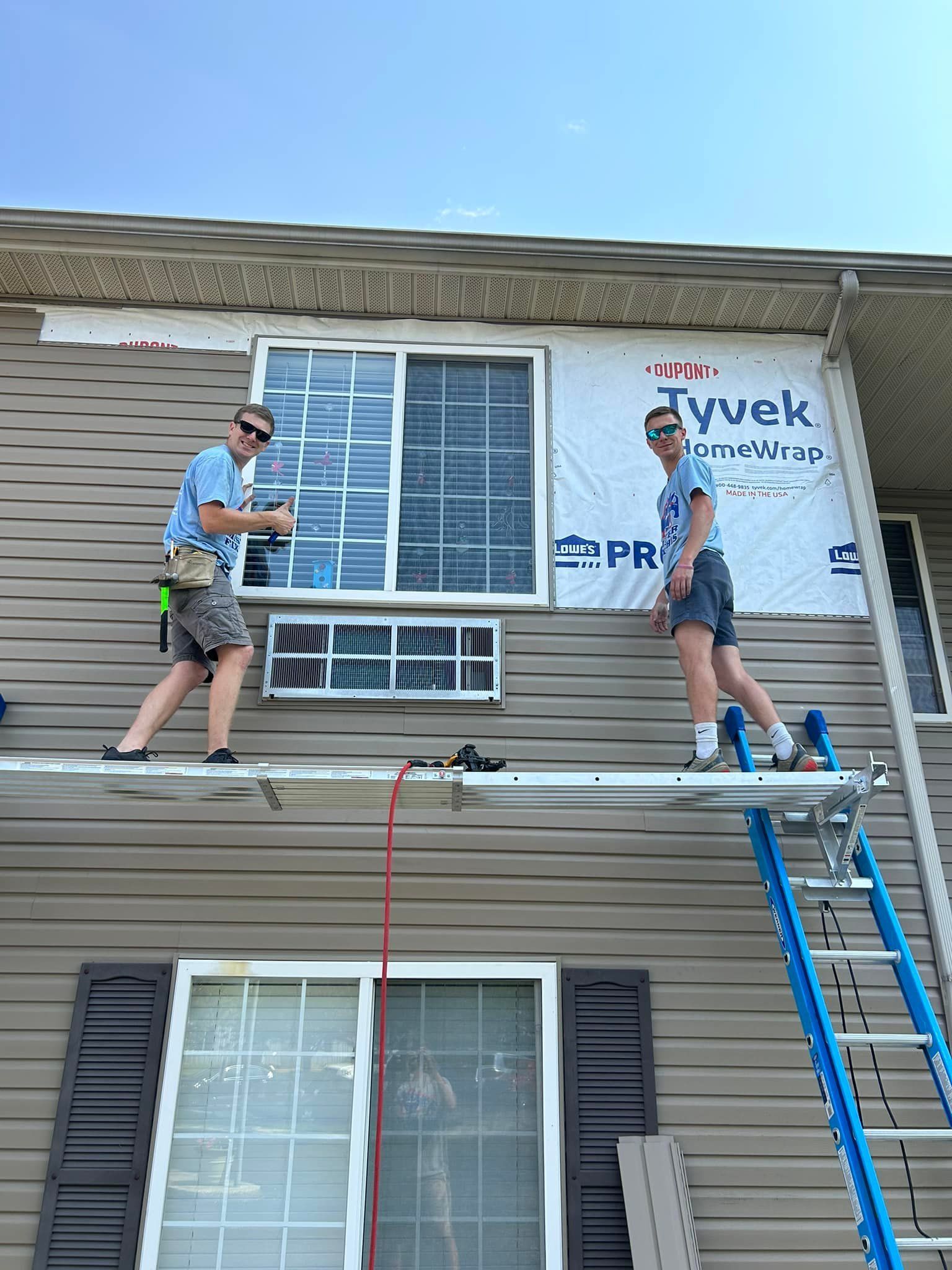Two people on a scaffolding installing siding on a house. One is holding a tool. Bright sunny day.