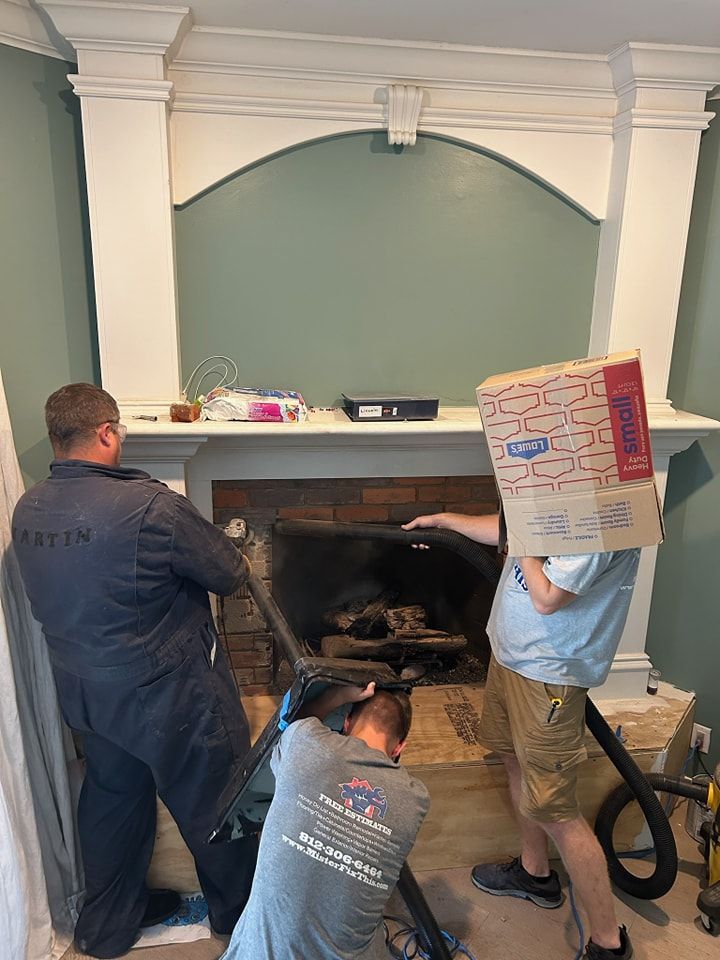 Three people cleaning a fireplace; one with a box on their head, two using vacuum hoses.
