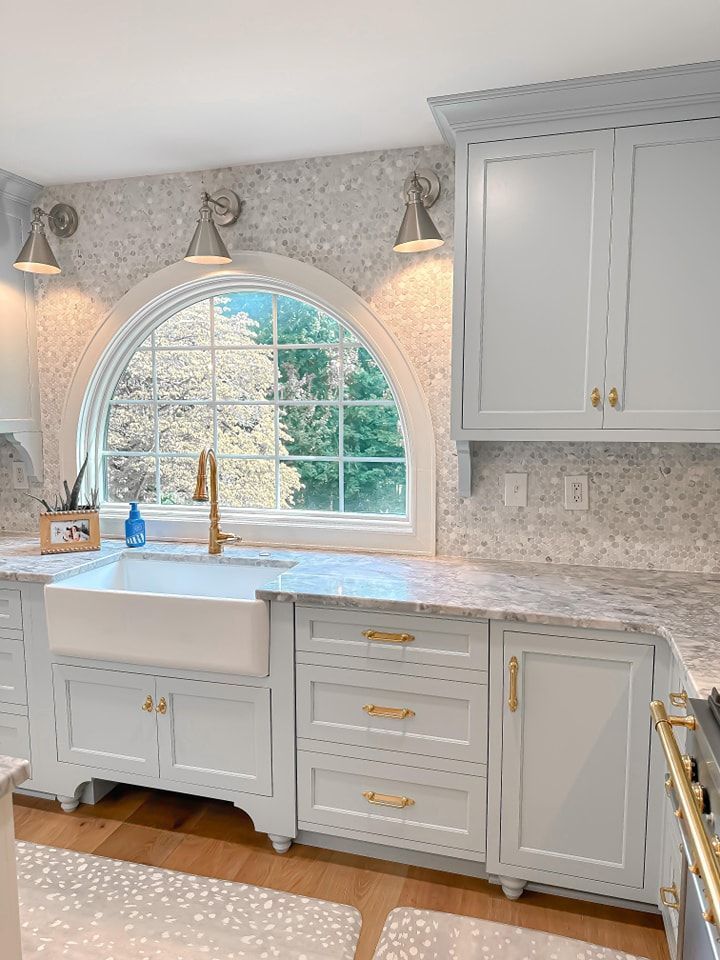 Light blue kitchen with arched window above a farmhouse sink, gold hardware, and mosaic tile backsplash.