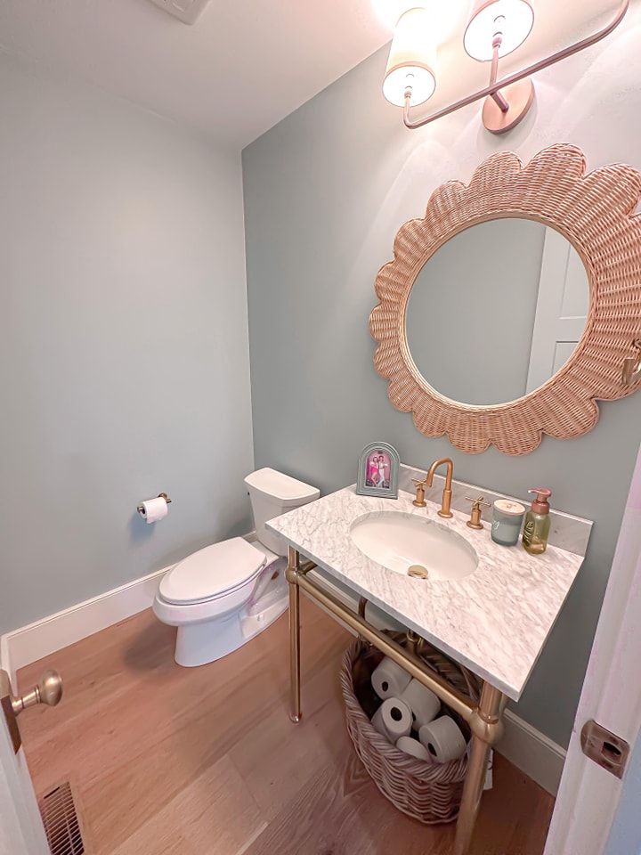 Powder room with light blue walls, wood floor, marble vanity, and a woven mirror.