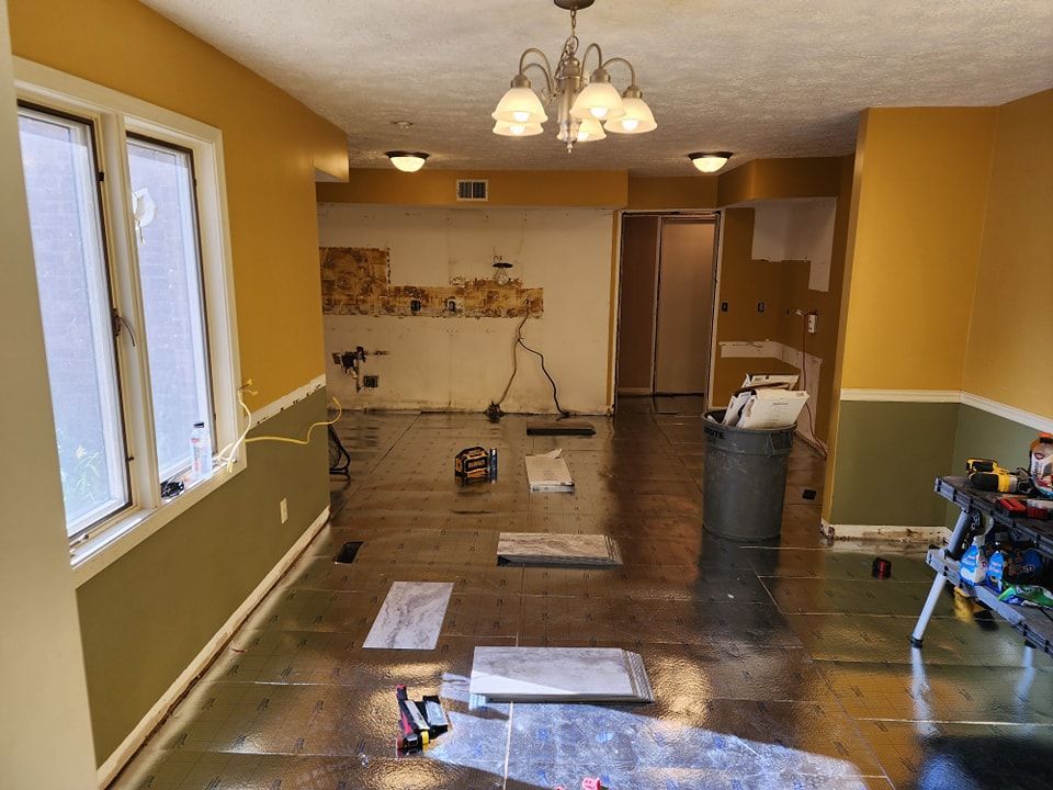 Room under renovation with exposed walls, tools, and wet flooring. Yellow and green walls, chandelier, and windows.