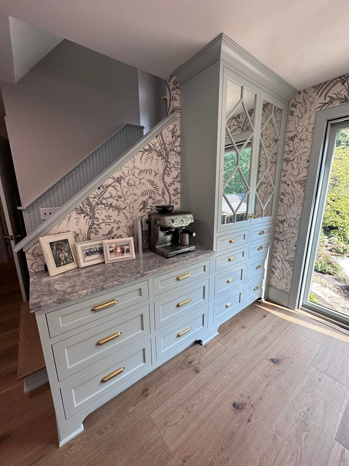 Light blue built-in cabinet with espresso machine, drawers, and glass-door upper cabinet, beside staircase with patterned wallpaper.