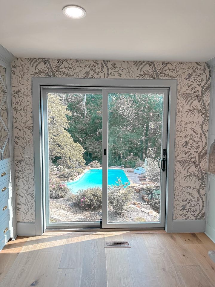 Sliding glass door frames pool view. Light-colored wallpaper surrounds, light blue trim, wooden floor.