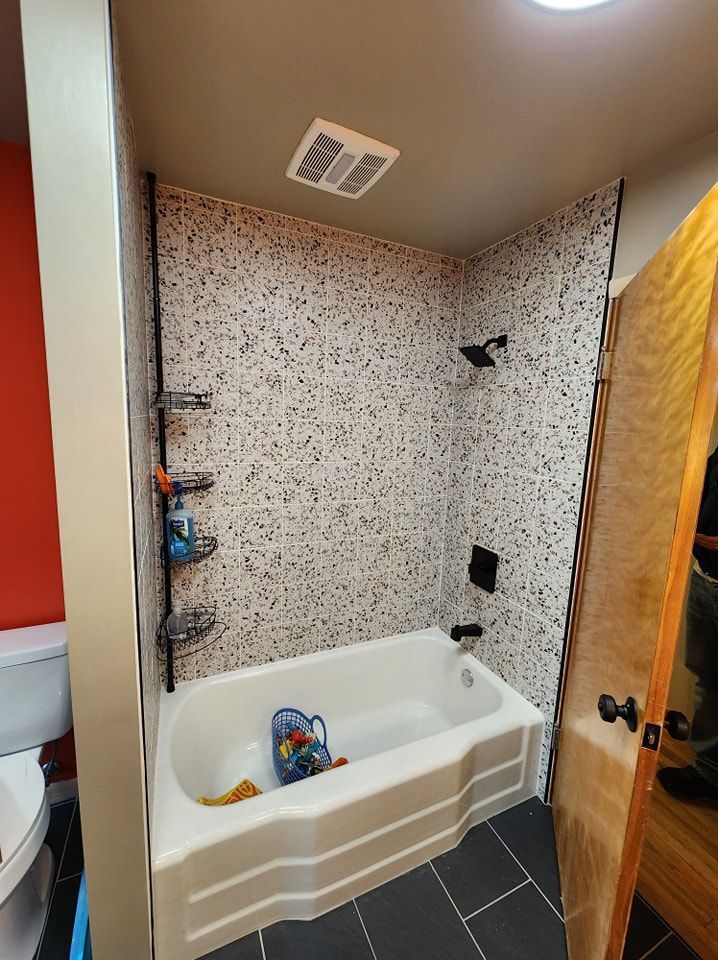 Small bathroom with a white bathtub and speckled tile walls. A showerhead and shelves are visible.