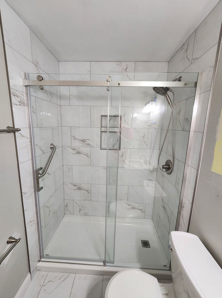 A modern, white-tiled shower with a sliding glass door, grab bar, and built-in niche. A toilet is to the right.