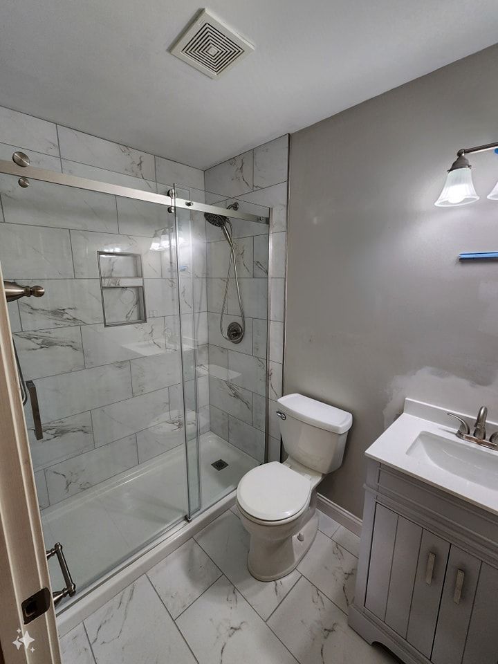 Bathroom with a glass shower, toilet, vanity, and marble-like tile flooring and shower walls.