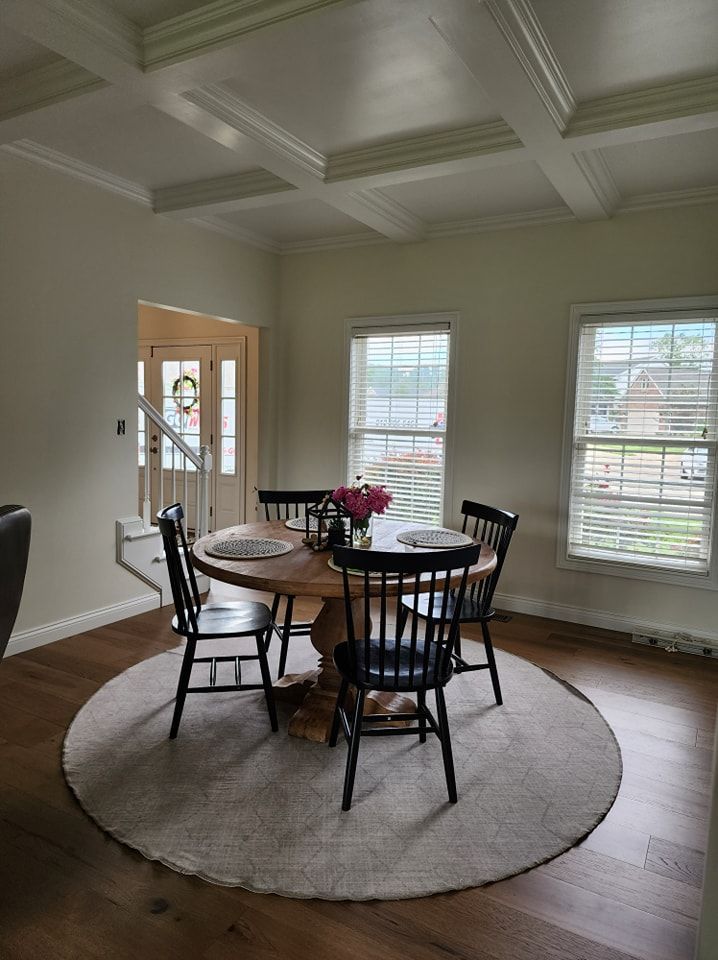 Dining room with round wooden table, four black chairs, rug, and windows.