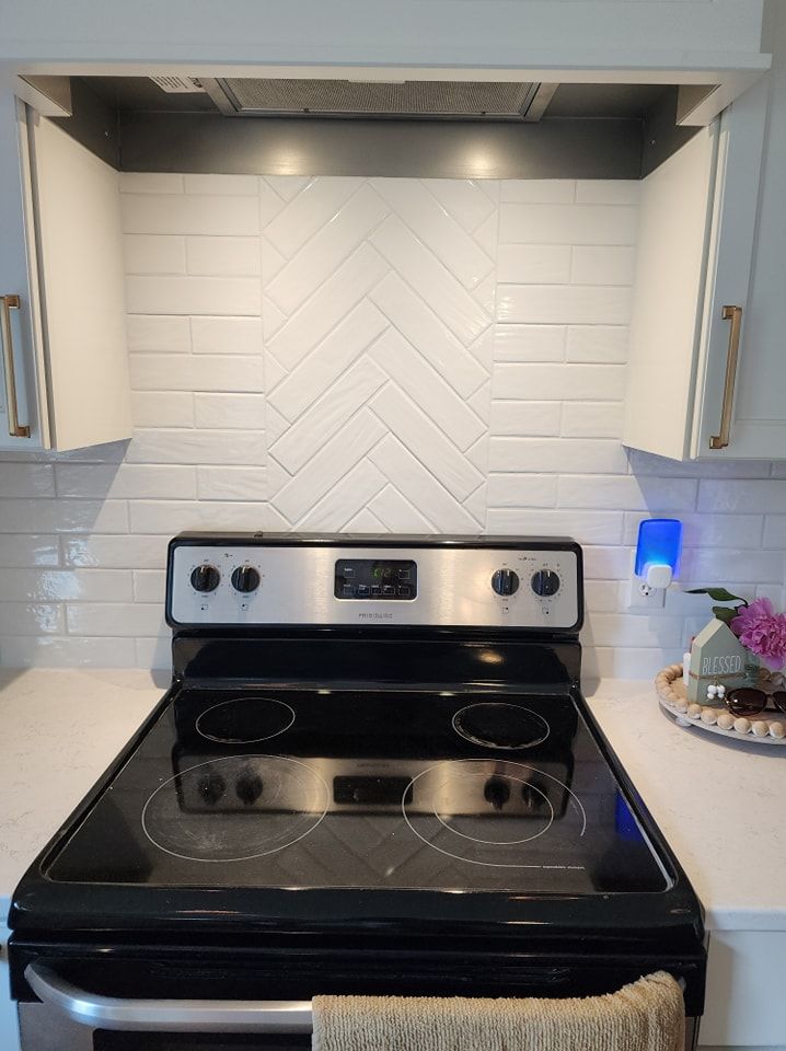 White kitchen backsplash with herringbone pattern, above a black stovetop, with cabinets on either side.