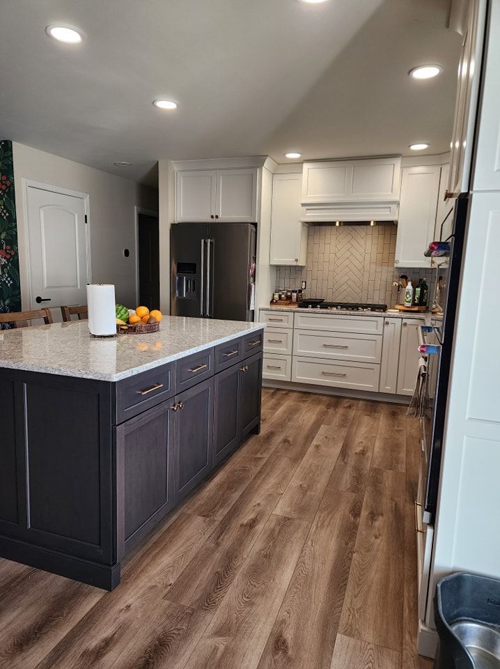 Modern kitchen with dark island and white cabinets, stainless steel fridge, and wood floors.