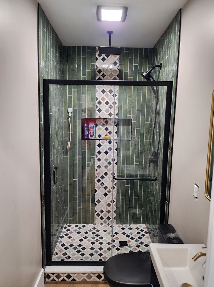 A modern, tiled shower with glass doors, a black frame, and green and patterned tiles. A small sink is on the right.