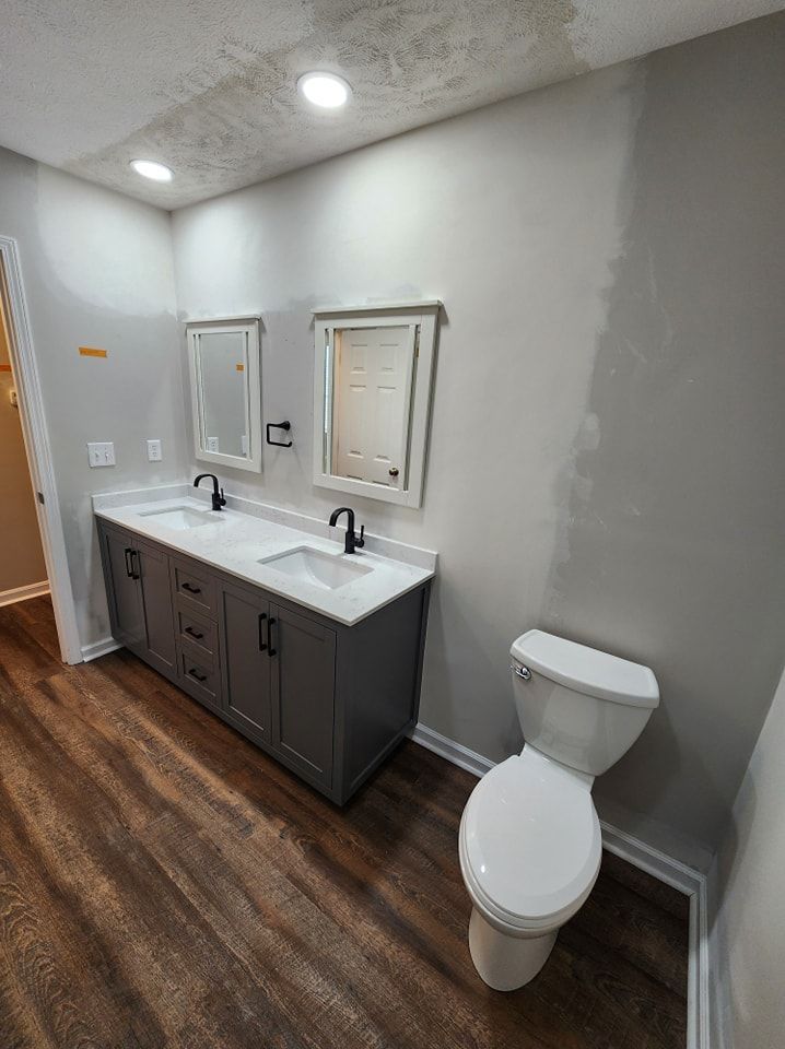Bathroom with gray vanity, white countertop, black faucets, and white toilet. Brown floor and light gray walls.