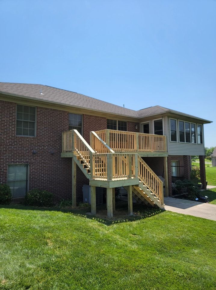 Wooden deck and stairs attached to a brick building, set against a sunny sky and green grass.