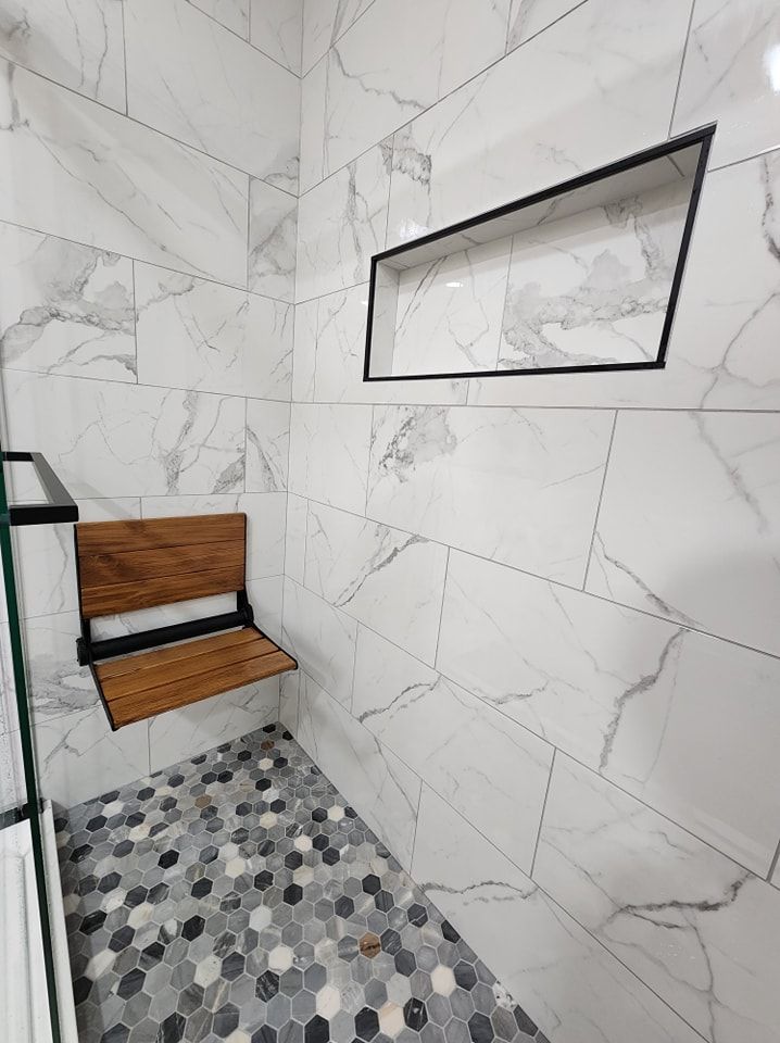 Shower with white marble tile, wood fold-down seat, and a built-in black-framed shelf. Hexagon tile floor.