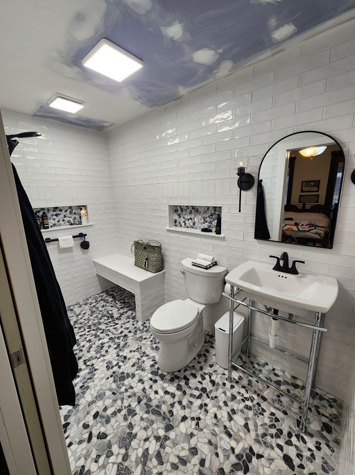 Bathroom with white subway tile walls, pebble floor, built-in bench, toilet, pedestal sink, and arched mirror.